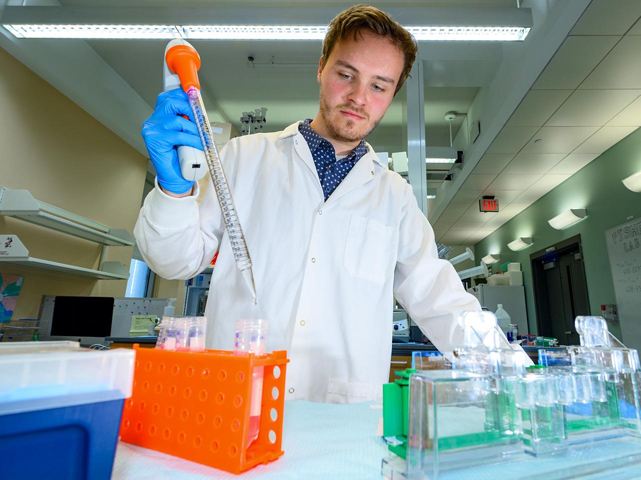Liam Babcock in a lab wearing a lab coat and gloves working with test tubes while using a pipette