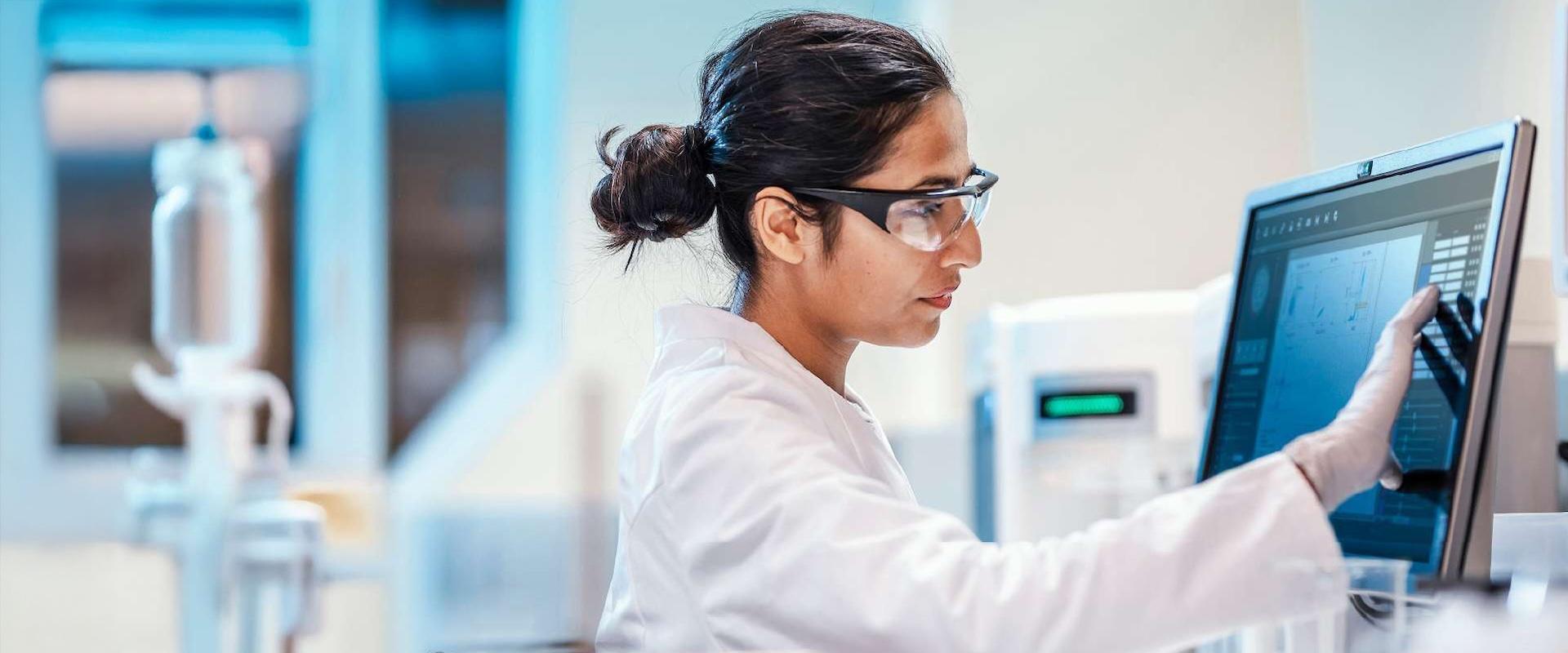 Scientist wearing a white coat and protective glasses review data on a computer monitor.
