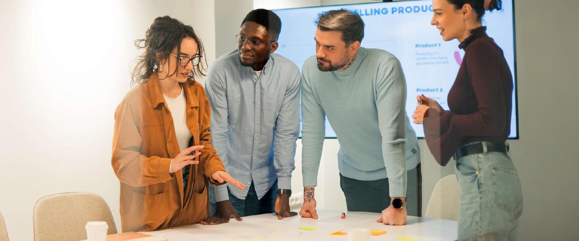 A diverse group of five people sitting around a small table talking about a pie chart in a presentation on a laptop.