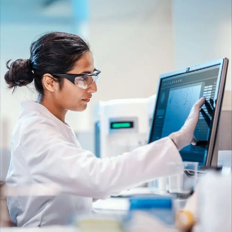 Scientist wearing a white coat and protective glasses review data on a computer monitor.