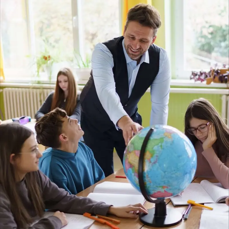 A teacher pointing to a point on the globe in front of a class of young people.