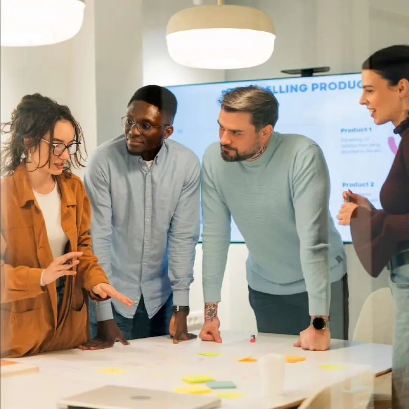A diverse group of five people sitting around a small table talking about a pie chart in a presentation on a laptop.
