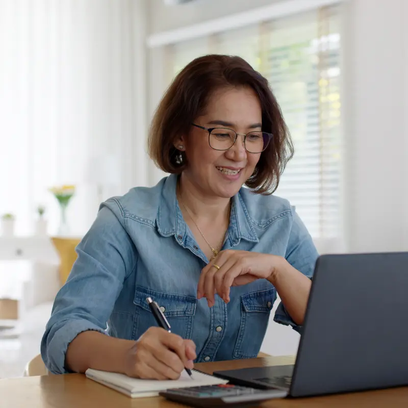 A student at her desk smiling while working in front of a laptop and writing on a notepad with a pen