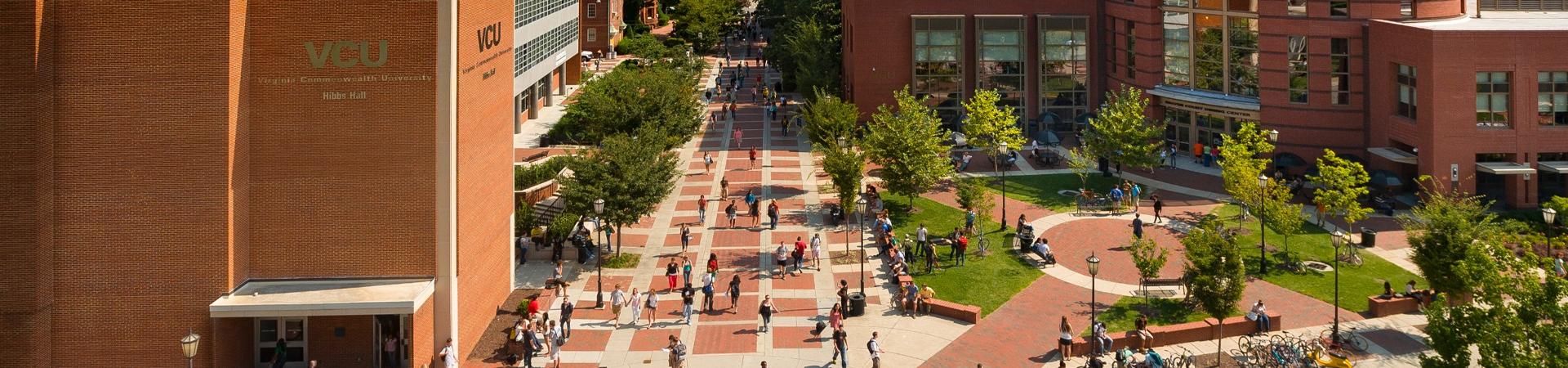 Daytime view of students walking across the Compass plaza on VCU’s Monroe Park Campus, surrounded by brick academic buildings, trees and outdoor seating.
