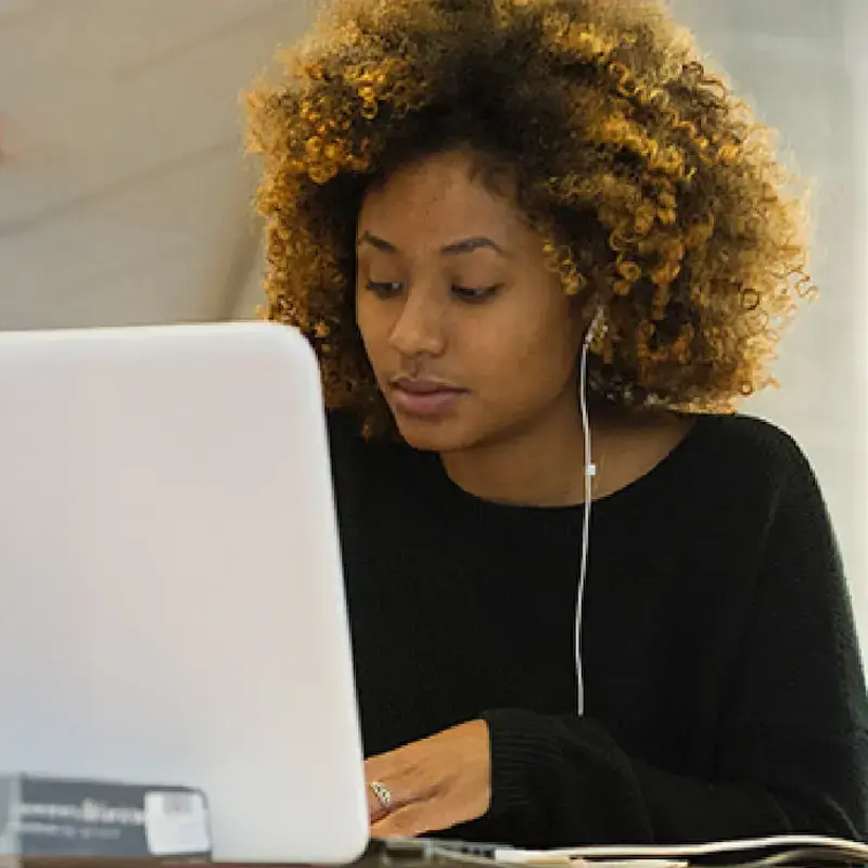Woman with curly hair wearing earbuds, working on a laptop in a modern indoor setting with soft lighting and blurred red foreground